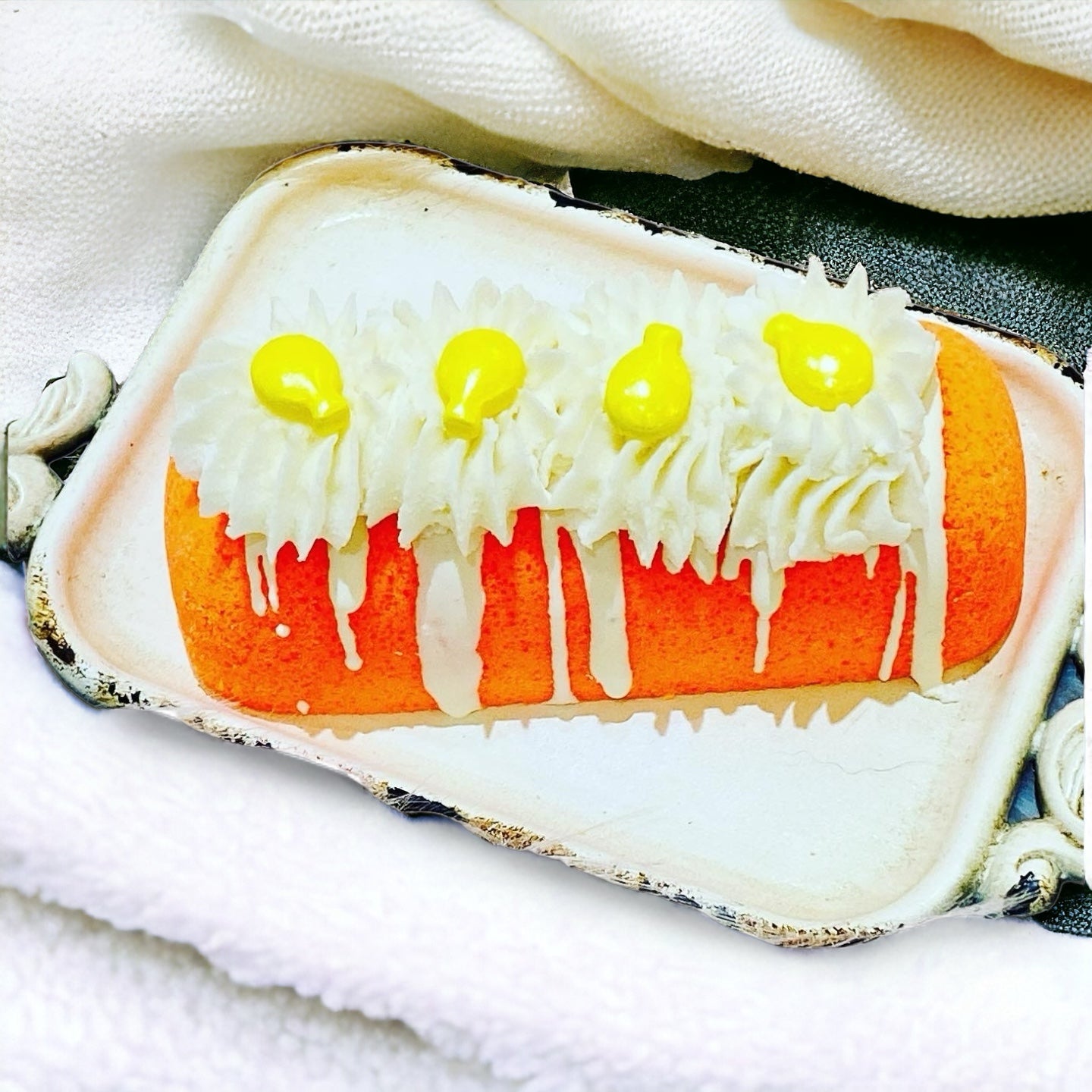 An orange twinkie bath bomb topped with white frosting and yellow sugar sprinkles on a white tray on top of a white towel