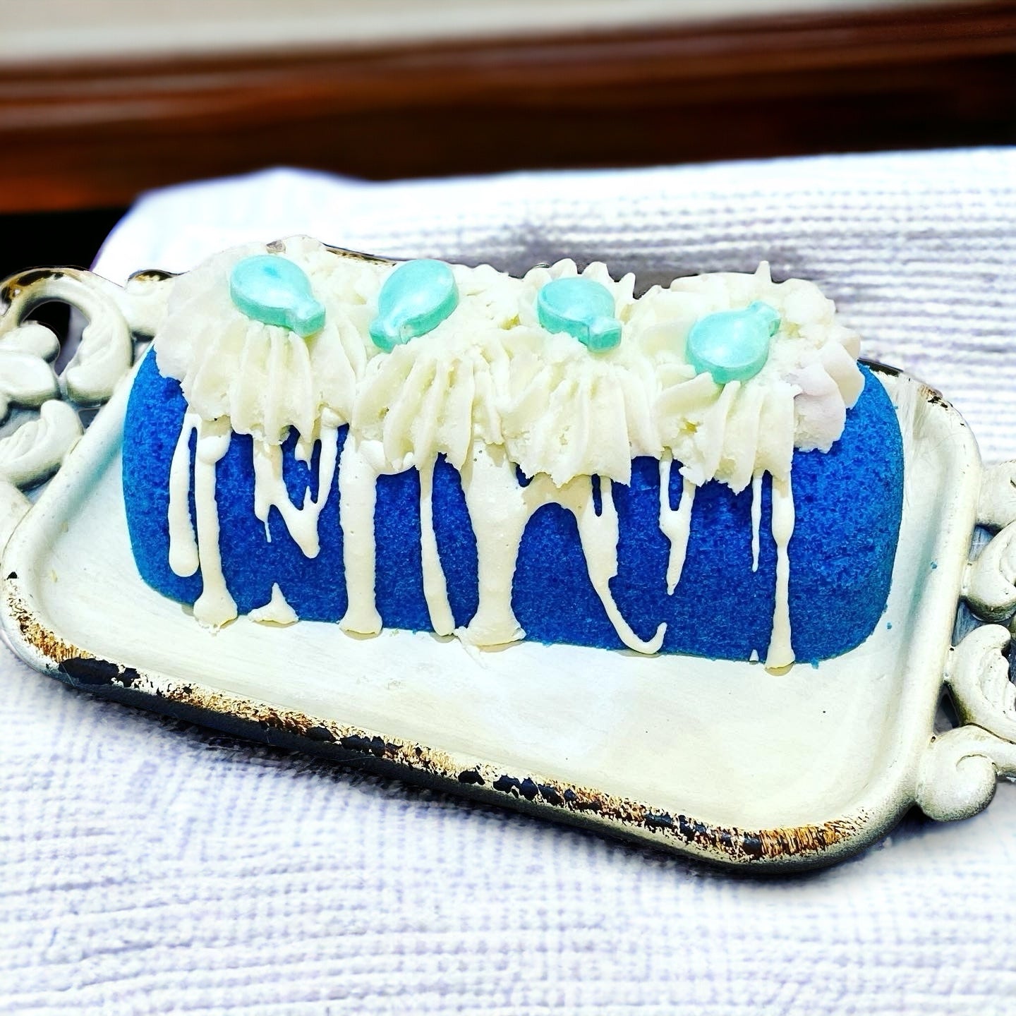 A blue twinkie bath bomb topped with white frosting and teal sugar sprinkles on a white tray on a white towel