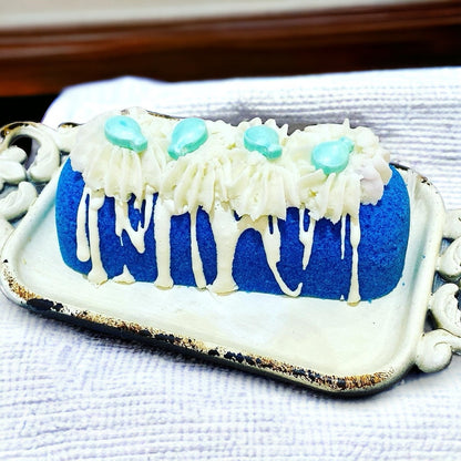 A blue twinkie bath bomb topped with white frosting and teal sugar sprinkles on a white tray on a white towel