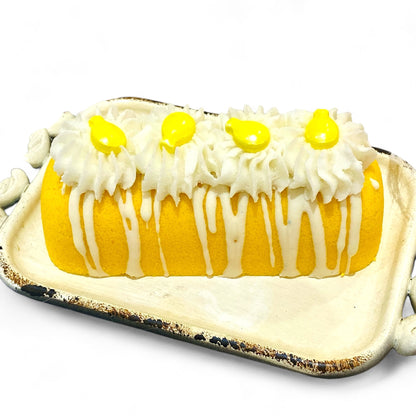 A yellow twinkie bath bomb topped with white frosting and yellow sugar sprinkles on a white tray against a white background