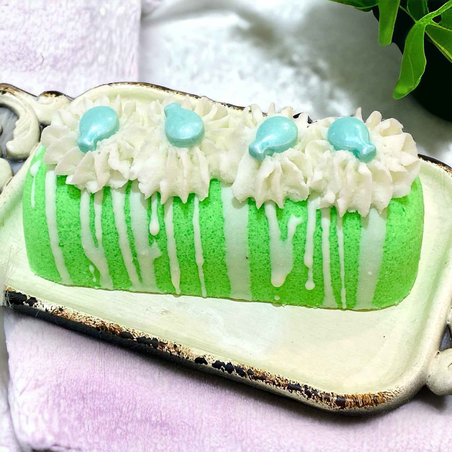 A green twinkie bath bomb topped with white frosting and teal sugar sprinkles on a white tray on top of a white towel