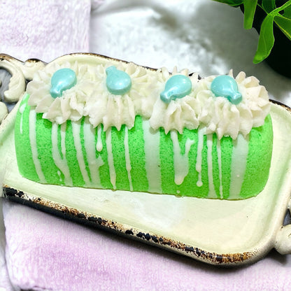 A green twinkie bath bomb topped with white frosting and teal sugar sprinkles on a white tray on top of a white towel