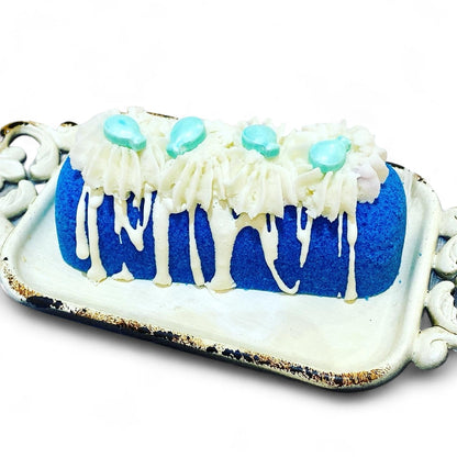 A blue twinkie bath bomb topped with white frosting and teal sugar sprinkles on a white tray against a white background