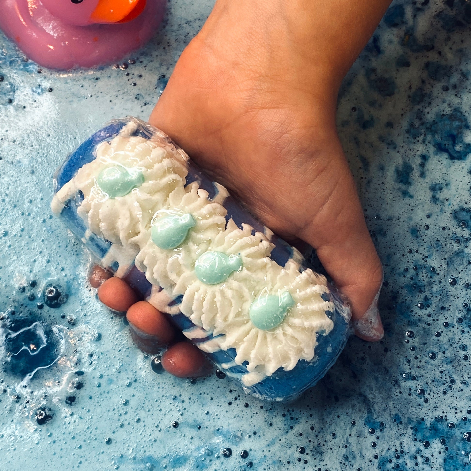 A blue twinkie bath bomb topped with white frosting and teal sugar sprinkles being held above blue water
